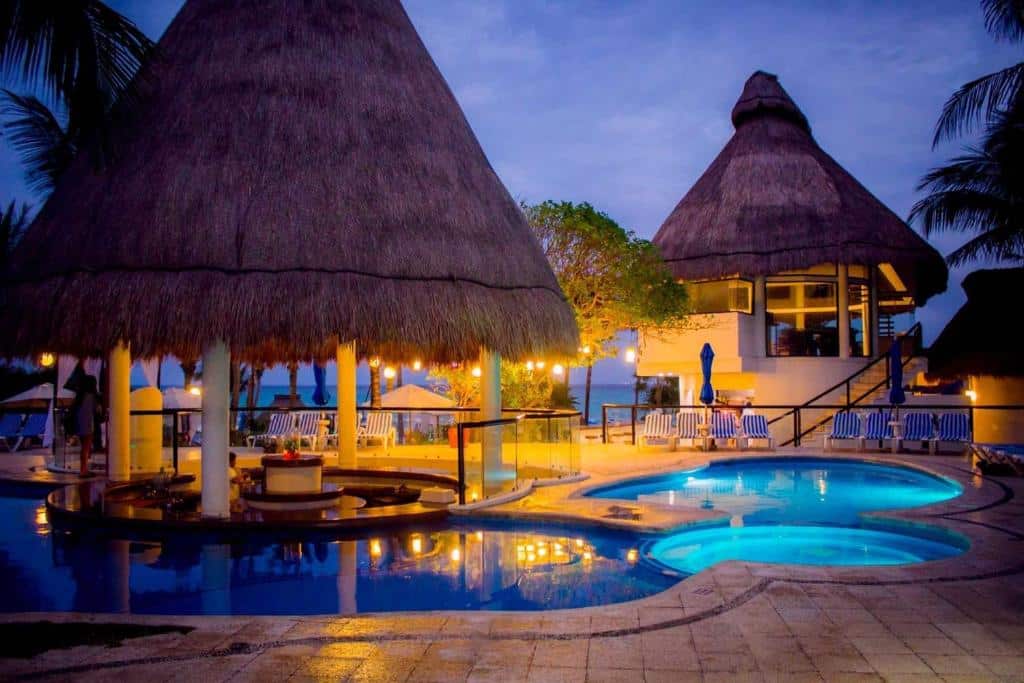 Piscina do The Reef Playacar Beach Resort & Spa, integrada com palapas de palha à noite, com luzes refletidas na água.