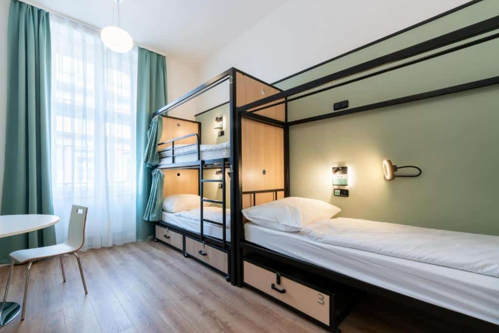 Quarto moderno do Luma Terra Prague Hostel, com dois beliches, chão de madeira clara, uma cadeira branca e uma mesa redonda junto a uma janela grande.