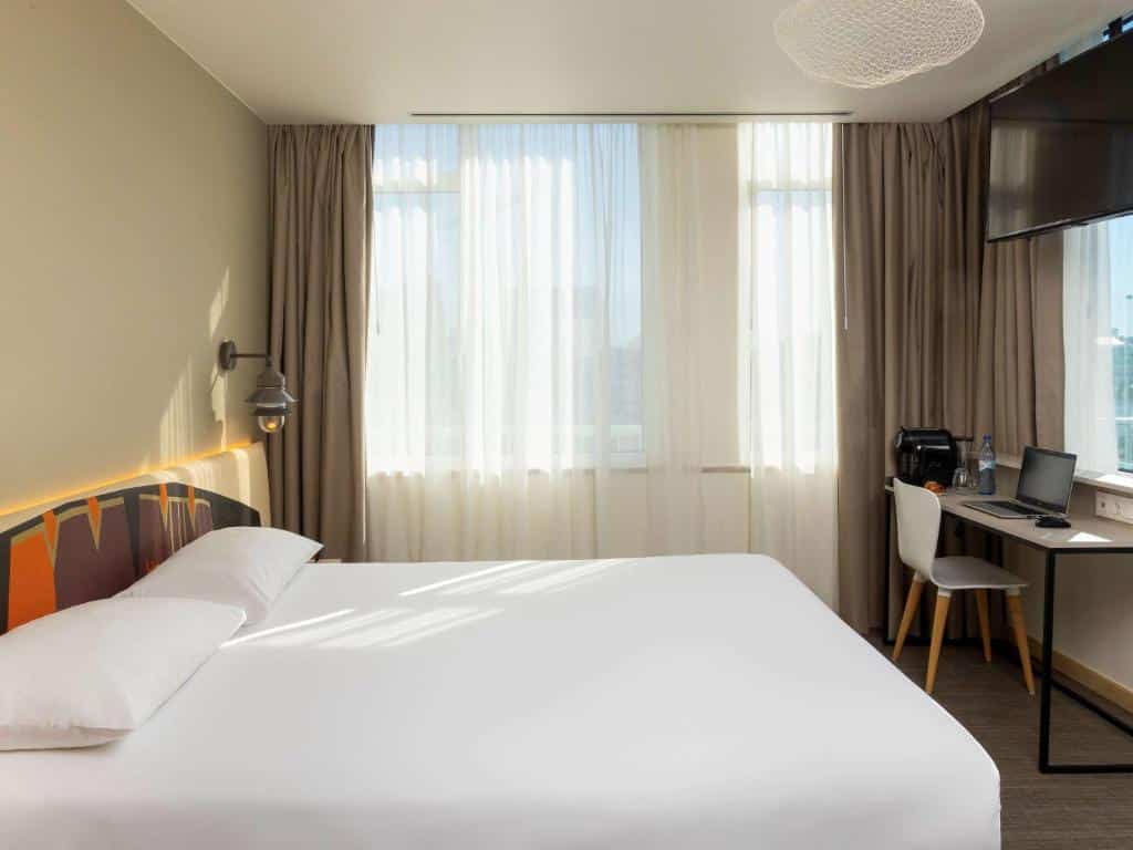 Quarto do Ibis Styles Lisboa Centro com cama grande, candeeiro de parede, secretária com cadeira, máquina de café e TV na parede.