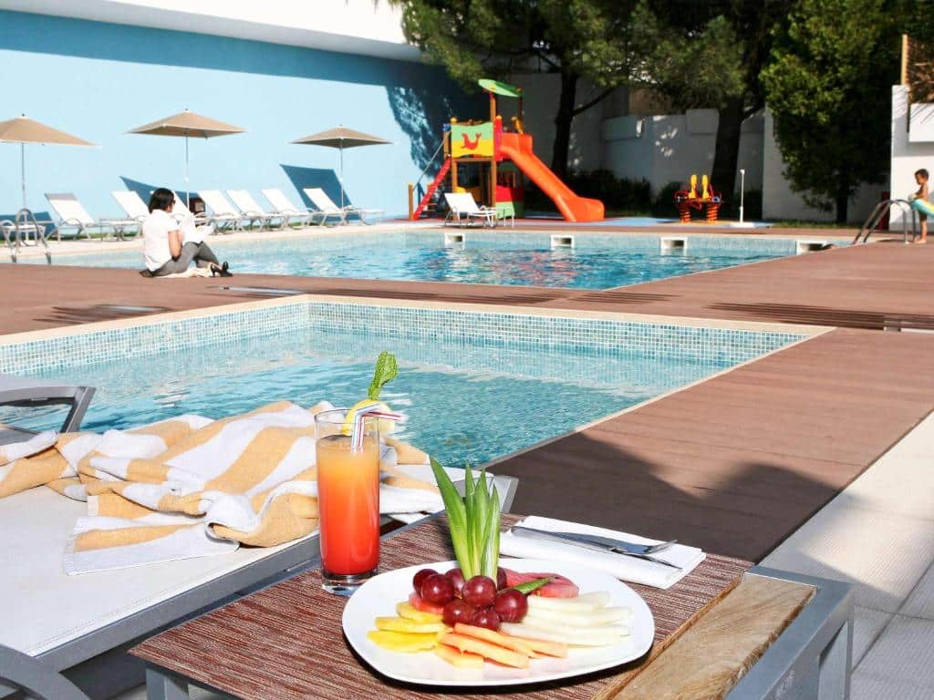 Zona junto à piscina do Novotel Lisboa, com fruta e uma bebida sobre uma mesa, espreguiçadeiras e guarda-sóis.