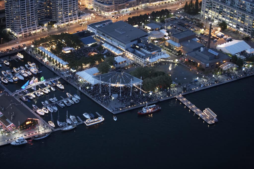 Embarcações e iates atracados numa marina iluminada, com centro de lazer e entretenimento ao fundo, incluindo grande estrutura coberta com teto em estilo de tendas