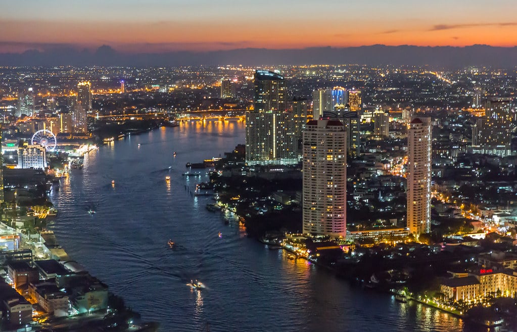 Vista aérea de Banguecoque ao anoitecer, com o rio, arranha-céus iluminados e as luzes da cidade sob um céu colorido.