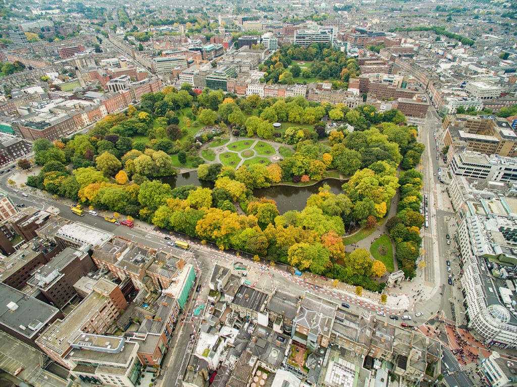 Vista aérea de um parque arborizado no centro da cidade, rodeado de edifícios. O parque tem jardins com flores, lagos e caminhos pedonais, entre árvores com folhagem de outono em tons de amarelo, laranja e verde.