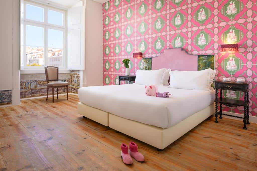 Quarto do Residentas Arco do Bandeira com uma cama de casal, uma janela grande, papel de parede azul e rosa, pavimento que imita madeira e mesas de cabeceira