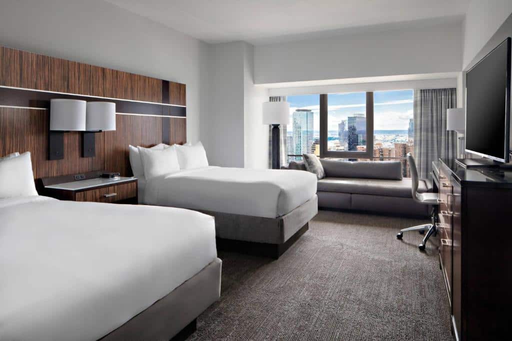No quarto do New York Marriott Marquis há duas camas de casal com cabeceiras de madeira escura. À direita, um sofá cinzento junto à janela, com vista para a cidade; à esquerda, uma secretária com cadeira de escritório.