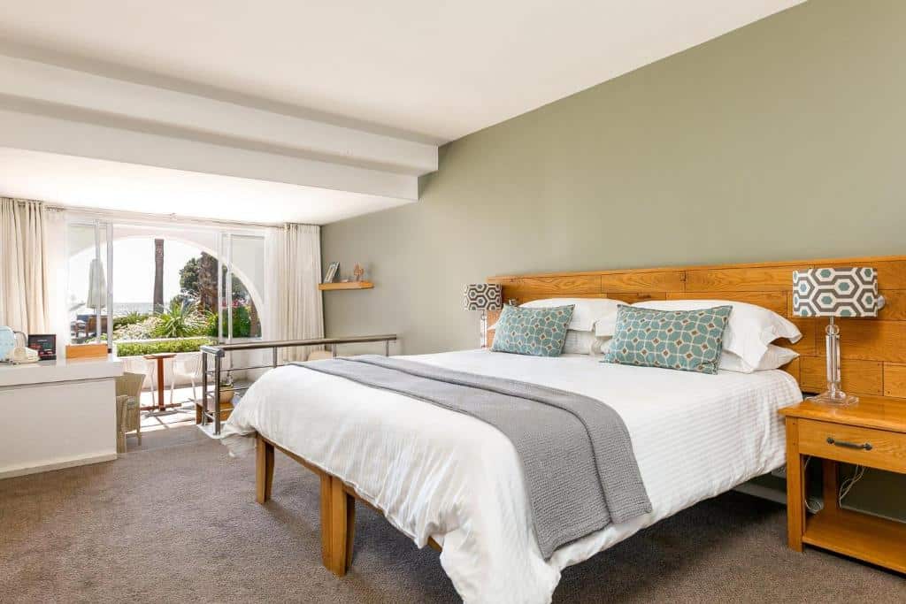 Quarto no The Bay Hotel com cama grande, cabeceira de madeira, duas mesas de cabeceira, candeeiros e janela com vista para um jardim.