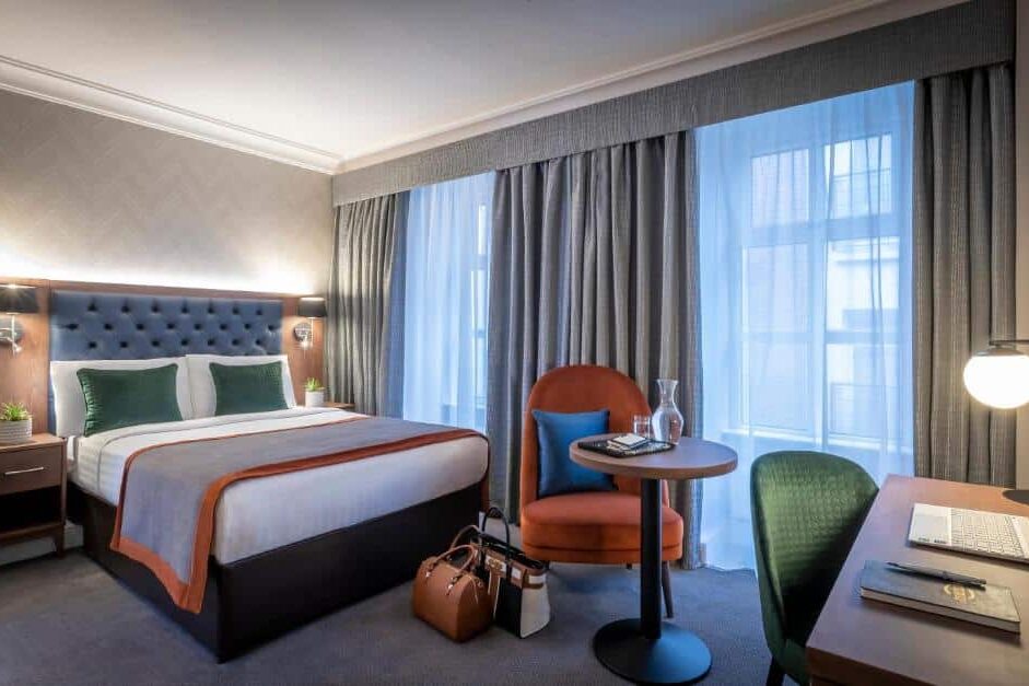 Quarto do The Fleet com cama de casal estofada em azul capitoné, mesa de canto com candeeiro e vaso de planta, cadeira redonda cor de laranja e secretária com portátil.