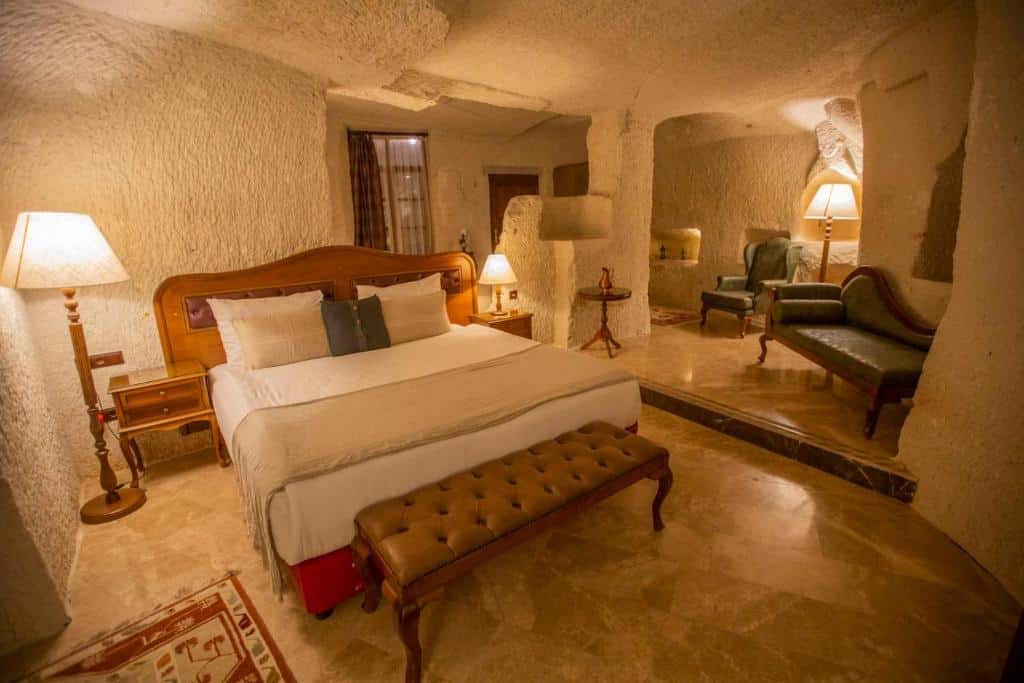 Quarto do Artemis Cave Suites & Spa, um dos hotéis da Capadócia, com paredes em textura de pedra, cama grande, candeeiros, poltronas e um banco de madeira aos pés da cama.