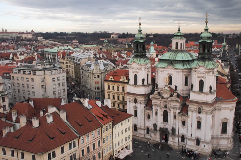 Vista aérea de uma cidade histórica europeia com edifícios de telhados vermelhos e uma grande igreja barroca sob um céu nublado.