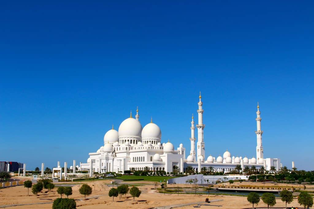 Mesquita Sheikh Zayed com cúpula branca e minaretes sob um céu azul claro, rodeada de vegetação.