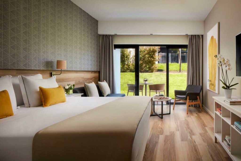 Quarto do Tivoli Alvor Algarve – All Inclusive Resort com cama de casal do lado esquerdo da imagem em frente a cama uma cômoda com TV presa na parede do lado esquerdo da imagem área de estar com sofá e cadeira. Representa hotéis all inclusive em Portugal.