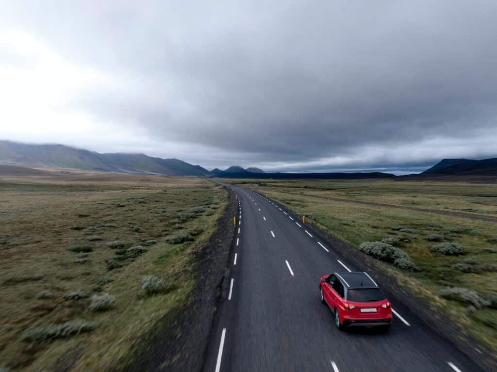 Carro vermelho com tejadilho preto numa estrada com campos verdes de ambos os lados, cadeia de montanhas ao fundo e nuvens densas a ocupar o céu.