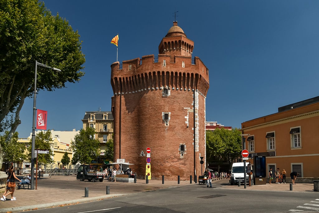 Torre circular de tijolo com ameias e uma bandeira no topo, numa praça de Perpignan rodeada de edifícios e árvores.
