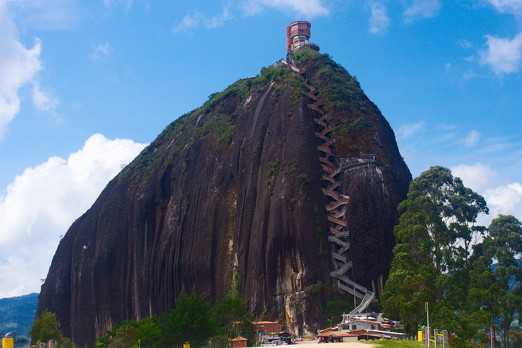 Grande formação rochosa de El Peñol, com uma escada em ziguezague a subir até uma estrutura no topo, rodeada de árvores sob um céu nublado.