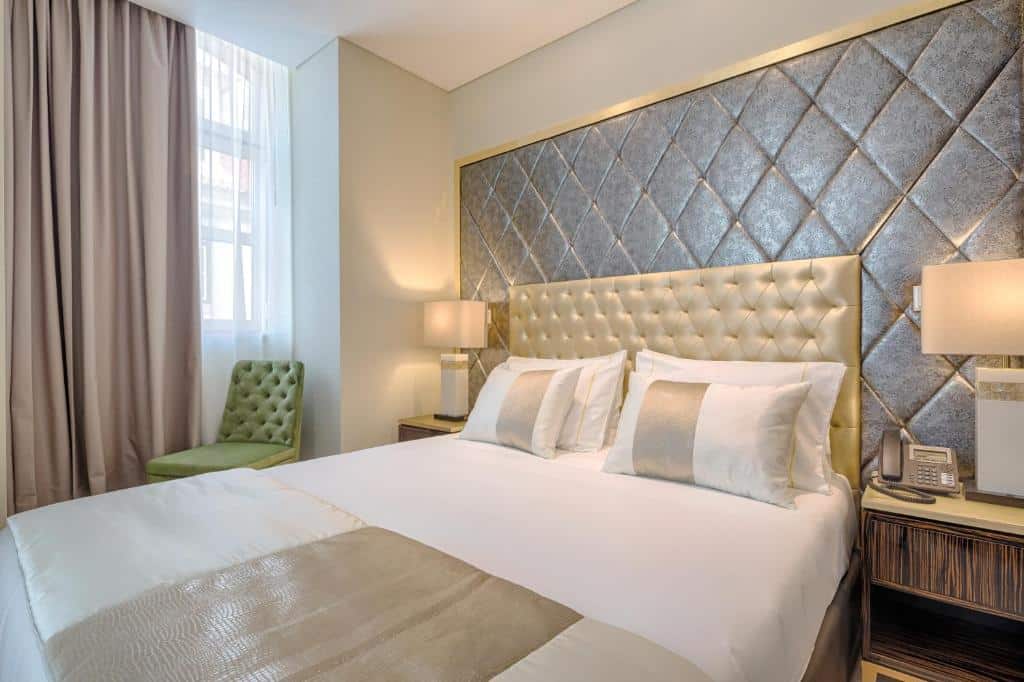 Quarto do Hotel MeraPrime Gold Lisboa com uma janela com cortinas e uma cadeira estofada por baixo, uma cama de casal com cabeceira estofada e duas mesas de cabeceira com candeeiros
