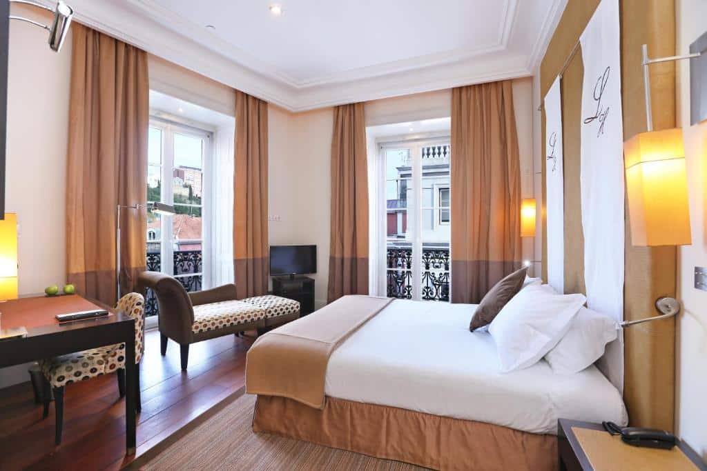 Quarto do Heritage Avenida Liberdade, com cama de casal e almofadas, duas varandas, uma poltrona, um móvel com televisão e uma pequena mesa com duas cadeiras estofadas