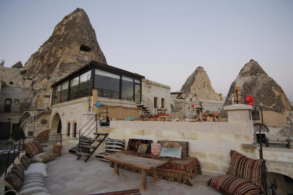 Terraço de pedra do Mithra Cave Hotel com almofadas e mesas estampadas, entre formações rochosas e edifícios de pedra ao anoitecer.