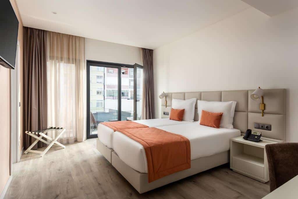 Quarto do Empire Marquês Hotel com uma pequena varanda com duas cadeiras, duas camas de solteiro com cabeceira estofada e candeeiros embutidos, duas mesas de cabeceira e uma televisão em frente às camas