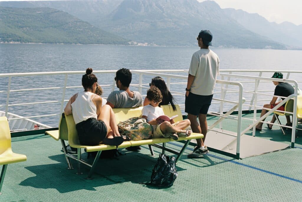 Pessoas sentadas num barco em Hvar a olhar para o mar e as montanhas. Uma criança lê um livro sentada e outra pessoa está de pé.