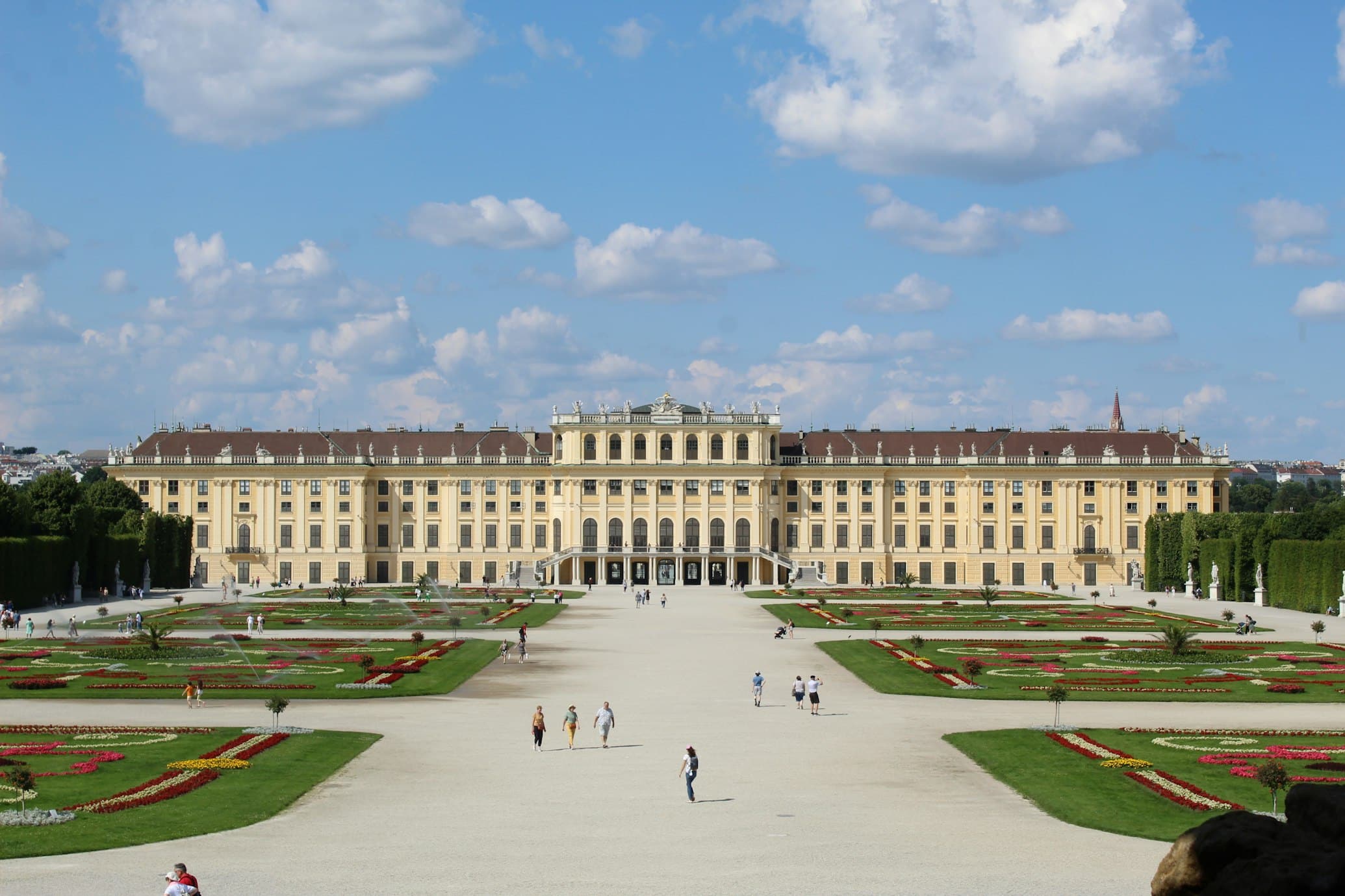 Palácio de Schönbrunn com os seus jardins e caminhos simétricos, sob um céu azul parcialmente nublado.