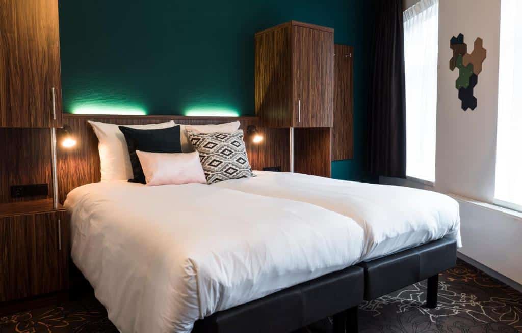 Quarto moderno do Linden Hotel, que ilustra onde ficar em Amesterdão: cama de casal, mobiliário de madeira escura, almofadas de destaque e luz natural de duas janelas.