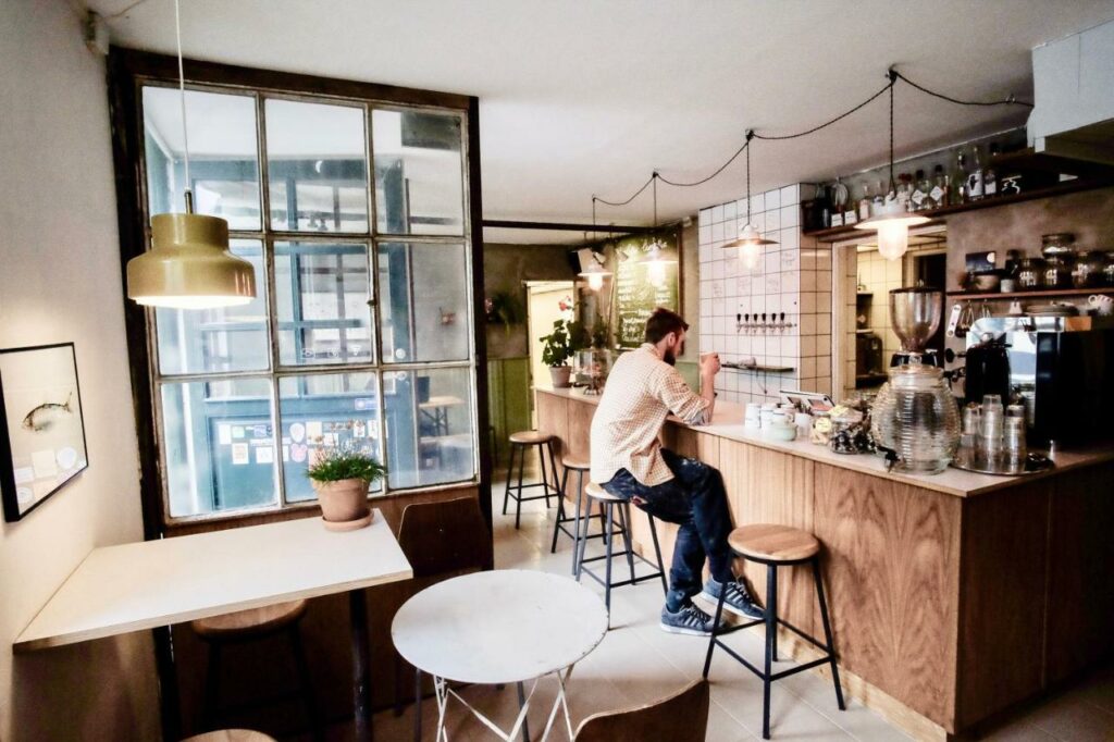 Cafetaria do Woodah Boutique Hostel. Há um balcão com uma pessoa sentada a beber café, uma mesa e várias decorações em redor.