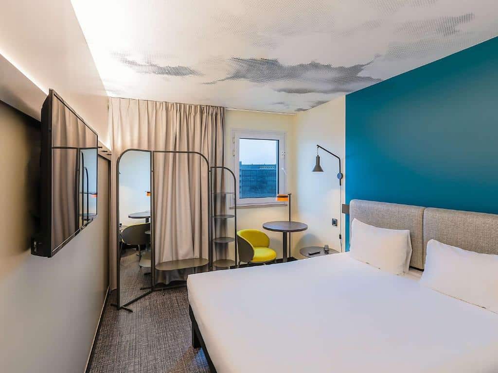 Quarto do Ibis Lisboa Alfragide com janela com cortinas, um bengaleiro integrado com um espelho de corpo inteiro, uma televisão e uma cama de casal