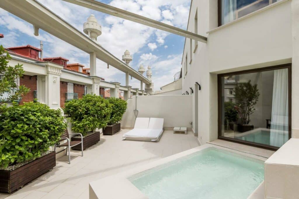 Terraço privativo com piscina do Room Mate Alicia. Espreguiçadeiras brancas estão junto a vasos de plantas dispostos à volta do local. Uma janela de vidro ao lado da piscina tem vista para o quarto.