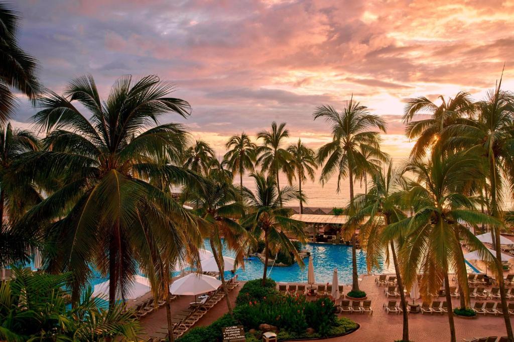 Palmeiras rodeiam uma piscina do Sheraton Buganvilias com espreguiçadeiras sob um céu colorido ao pôr do sol à beira-mar.