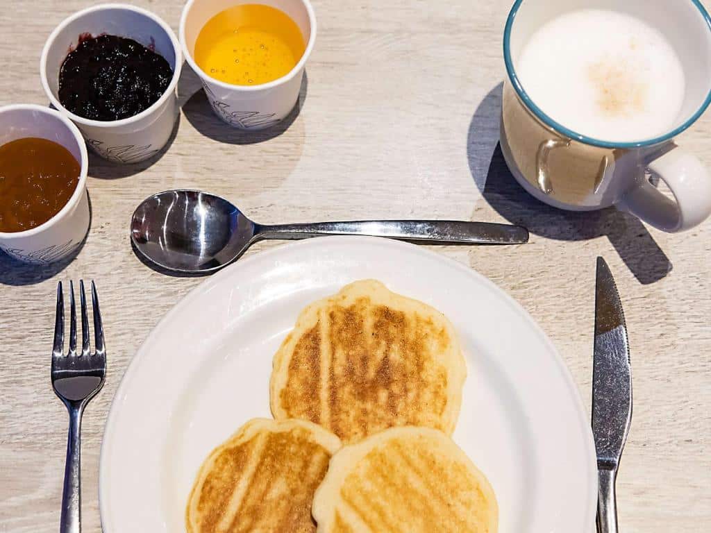 Prato com três panquecas, talheres, três taças de compota e uma caneca de leite com espuma sobre uma mesa.