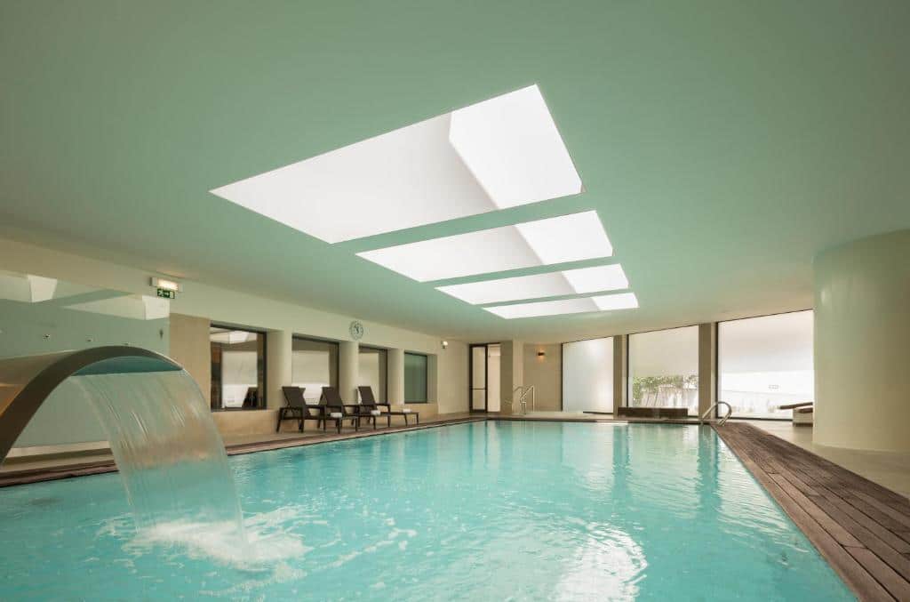 Piscina interior com jogo de água, espreguiçadeiras, deck de madeira e grandes clarabóias no teto, no Hotel Dom Gonçalo & Spa.