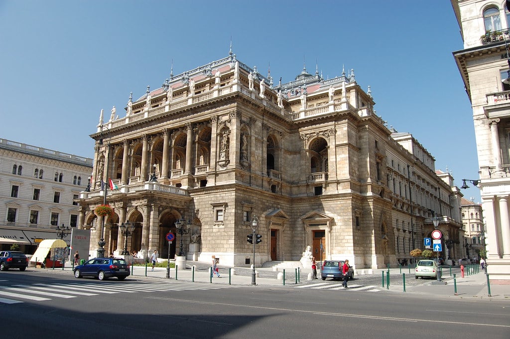 Um grande edifício de pedra ornamentado da Ópera Nacional da Hungria, com colunas e varandas, numa esquina da cidade sob um céu azul claro.