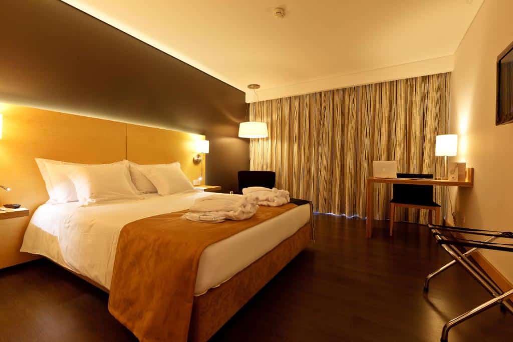 Quarto moderno do Hotel Mercure Braga Centro, com cama de casal, mesas de cabeceira, secretária com portátil, cadeira e janela ampla.
