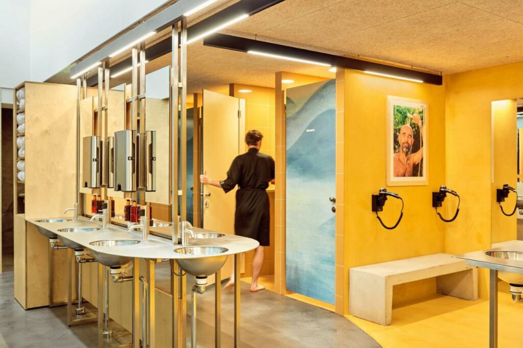 Casas de banho do CityHub Copenhagen. Vêem-se várias cabines separadas, uma zona com secadores de cabelo e uma área partilhada com lavatórios e espelhos.