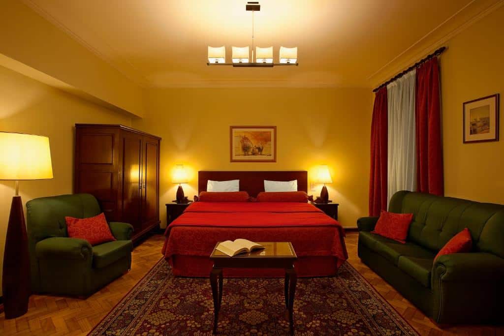 Quarto do Pão de Açúcar Hotel com cama de casal ao centro, duas cómodas ao lado, um roupeiro e uma pequena sala de estar com dois sofás verdes e uma mesa de madeira.