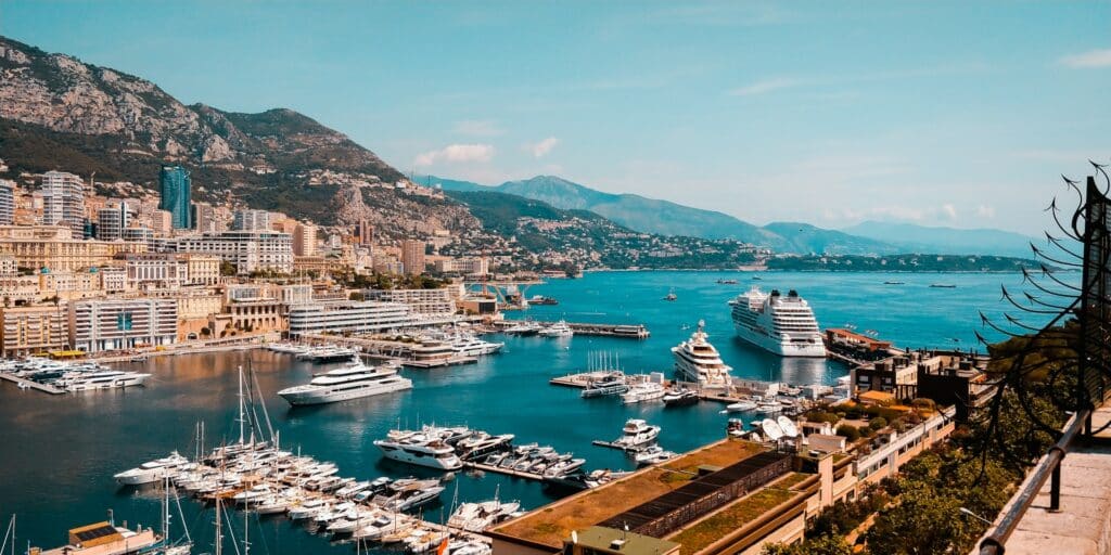 Monte Carlo, no Mónaco, com um cais repleto de embarcações de luxo num mar azul e montanhas com alojamentos sofisticados.