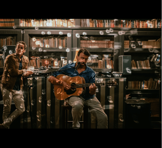 Dois homens a atuar numa sala de estilo biblioteca na Casa do Livro, com estantes de livros ao fundo. Um toca guitarra e canta. Representa o que fazer à noite no Porto.
