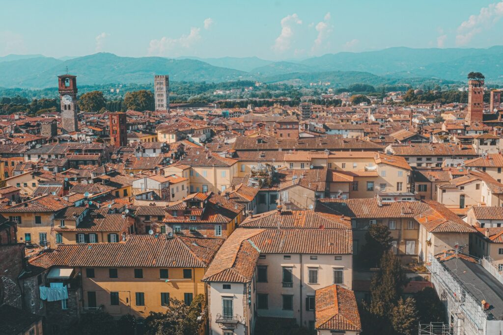 Vista aérea de Lucca, na Itália, com um mar de telhados de terracota e antigas torres medievais a elevarem-se acima das construções. A cidade está rodeada por montanhas ao fundo. As torres históricas e os edifícios oferecem um vislumbre da arquitetura e do planeamento urbano desta região da Toscana.