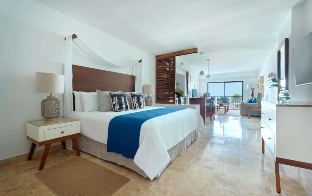 Quarto de hotel moderno com uma cama grande, detalhes em azul, pavimento de mármore, zona de estar e uma janela com vista para o mar.