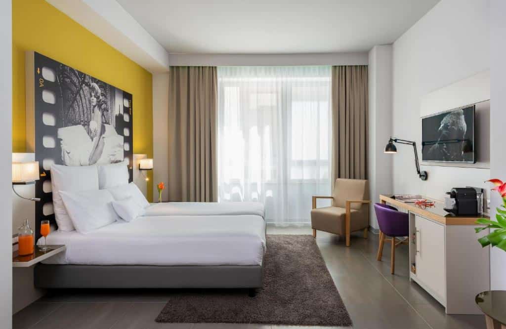 Quarto com cama e janela amplas, televisão, parede e poltrona amarelas no NYX Hotel Milan by Leonardo Hotels