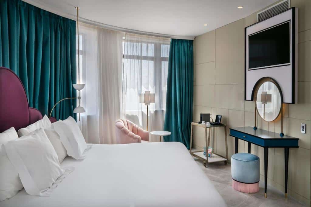 Quarto do Vincci Capitol, um dos melhores hotéis em Madrid. Decorado em tons de azul e rosa, tem uma cama de casal com roupa de cama branca em frente a um toucador com banco e espelho, e uma TV na parede por cima. Ao lado fica um aparador com comodidades para chá e café. As janelas ocupam as paredes dos lados da cama e uma poltrona com mesinha e candeeiro fica em frente ao aparador. Há cortinas ciano-escuro e brancas diáfanas nas janelas.