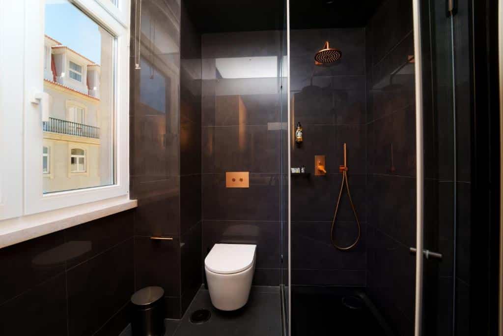 Casa de banho do Golden Lisbon Hotel com cabina de duche em vidro, sanita e uma janela branca, com revestimento em porcelanato preto