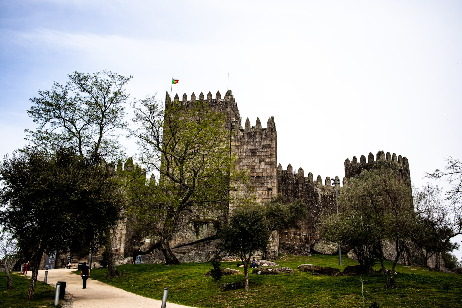 Castelo de Guimarães em pedra, com torres ameadas e uma bandeira no topo, rodeado de árvores e um caminho em primeiro plano.