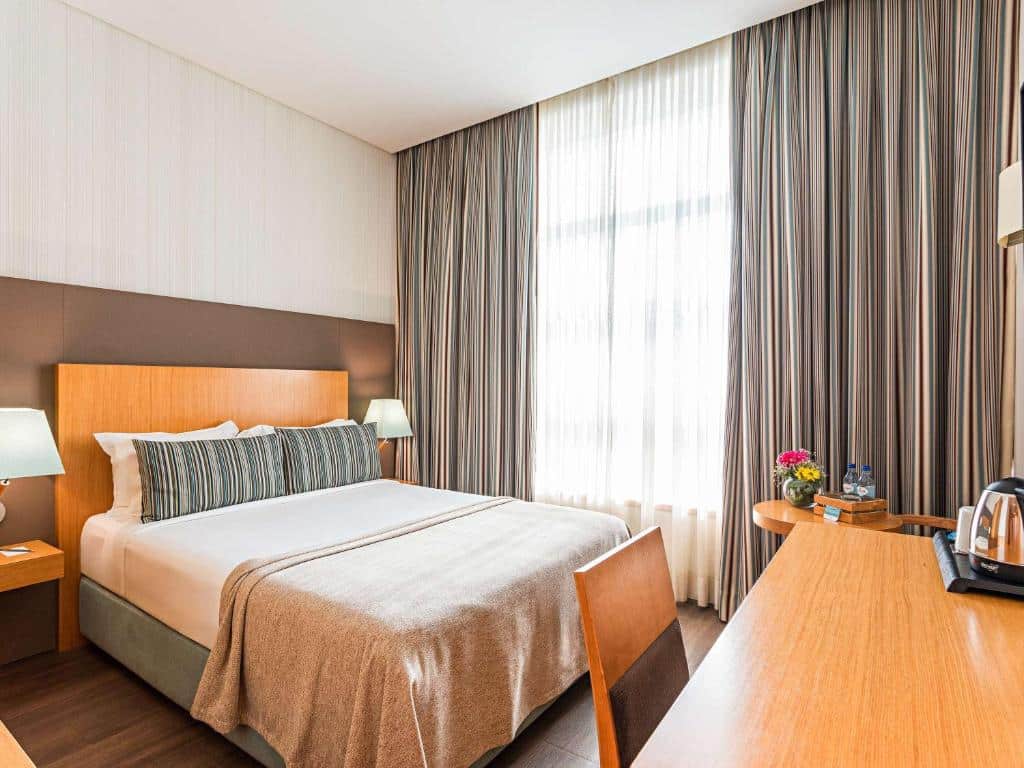 Quarto do Mercure Lisboa Almada com cama de casal arrumada, candeeiros de cabeceira, secretária de madeira com chaleira e janela com cortinas.