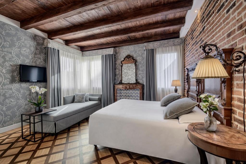 Quarto de hotel elegante com teto de vigas de madeira, paredes de tijolo à vista, cama de casal com cabeceira, sofá cinzento, candeeiro de pé antigo e móveis de madeira rústicos