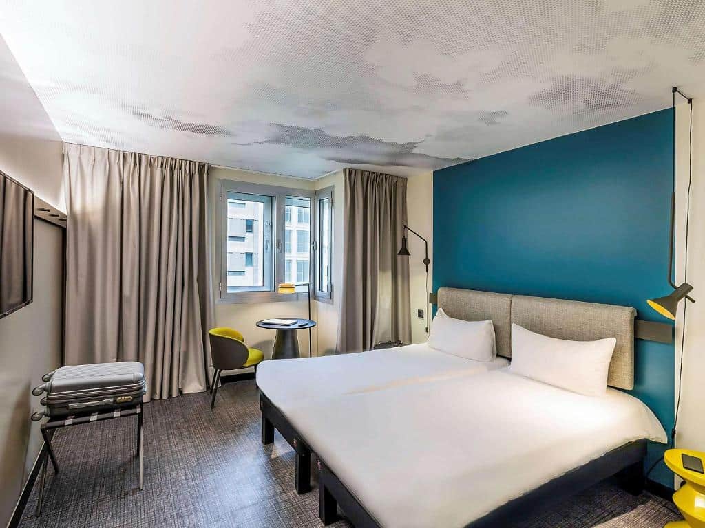 Quarto do Hotel Ibis Lisboa Saldanha com duas camas, parede com detalhes em azul-petróleo, duas janelas com cortinas e mesa com cadeiras verdes.