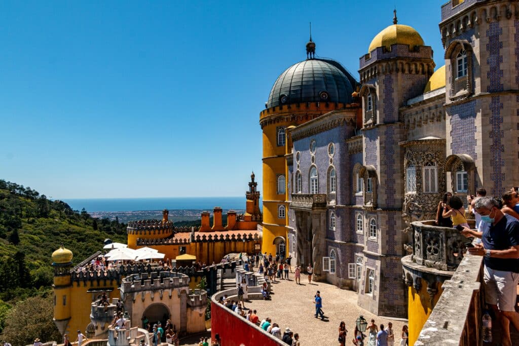 Grande construção com cúpulas e torres, fachada com azulejos azuis e uma área amarela, com várias pessoas a caminhar e a tirar fotografias.