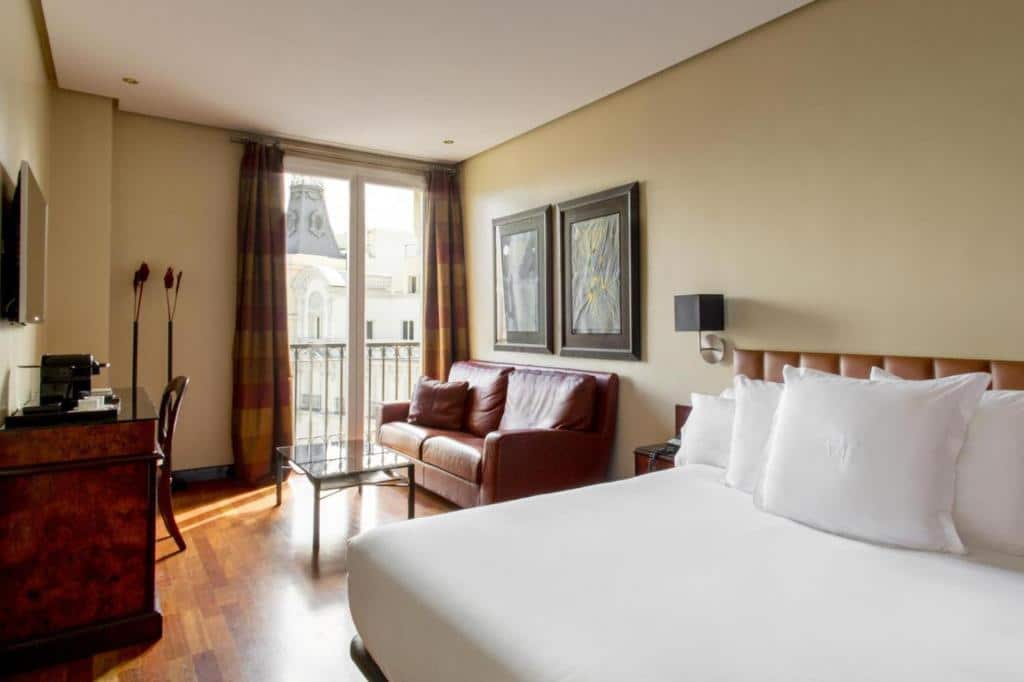 Quarto do Hotel Villa Real, um dos melhores hotéis em Madrid. A cama de casal tem roupa de cama branca e mesinha de cabeceira com candeeiro dos dois lados. Ao lado fica uma zona de estar com sofá e mesinha, virada para a secretária com comodidades para fazer café, cadeira e uma TV. Ao fundo fica uma grande janela com cortinas e vista para a cidade. Há dois quadros na parede por cima do sofá.
