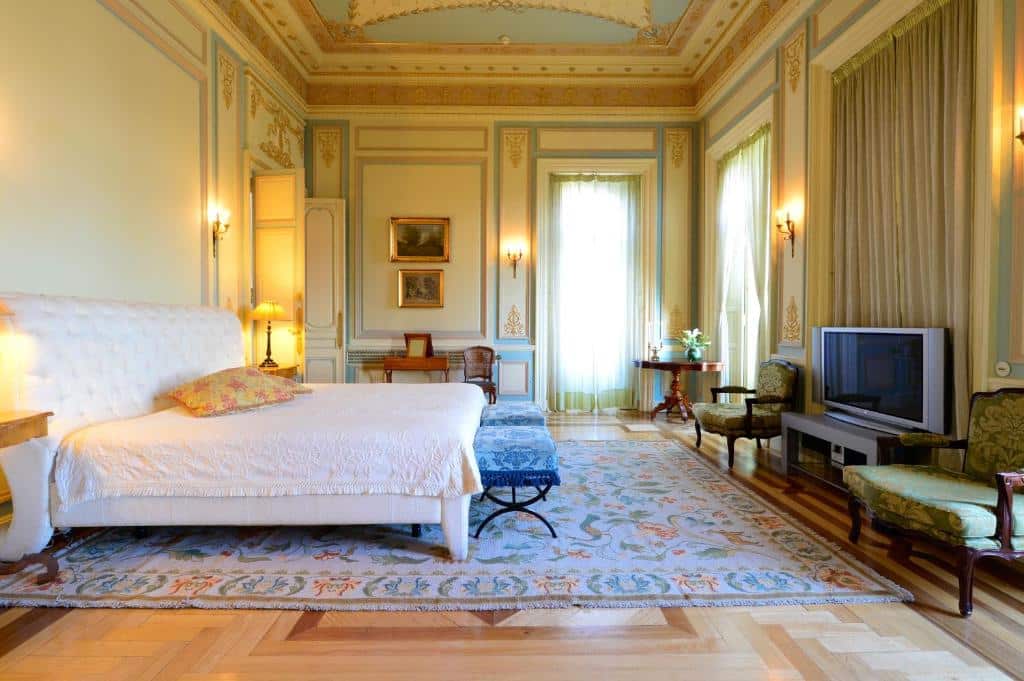 Quarto espaçoso do Pestana Palace Lisboa com três janelas, cama de casal, tapete amplo, poltrona e teto alto com decoração dourada