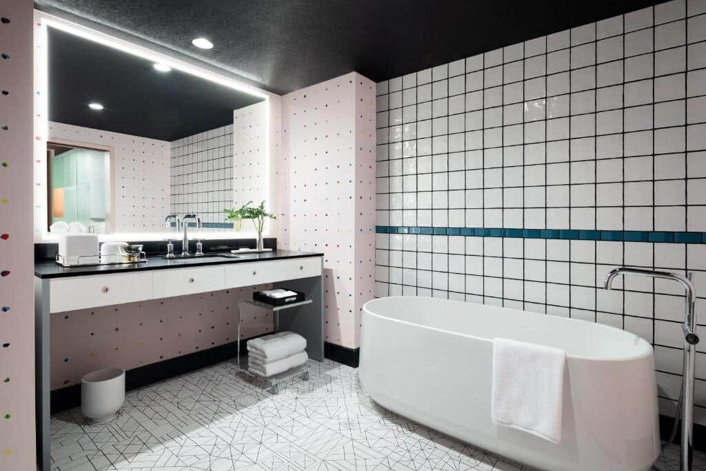 Casa de banho do Graduate New York com uma banheira branca à direita. À esquerda, um lavatório preto com duas cubas e um espelho grande. A parede é decorada com azulejos brancos e pormenores coloridos.